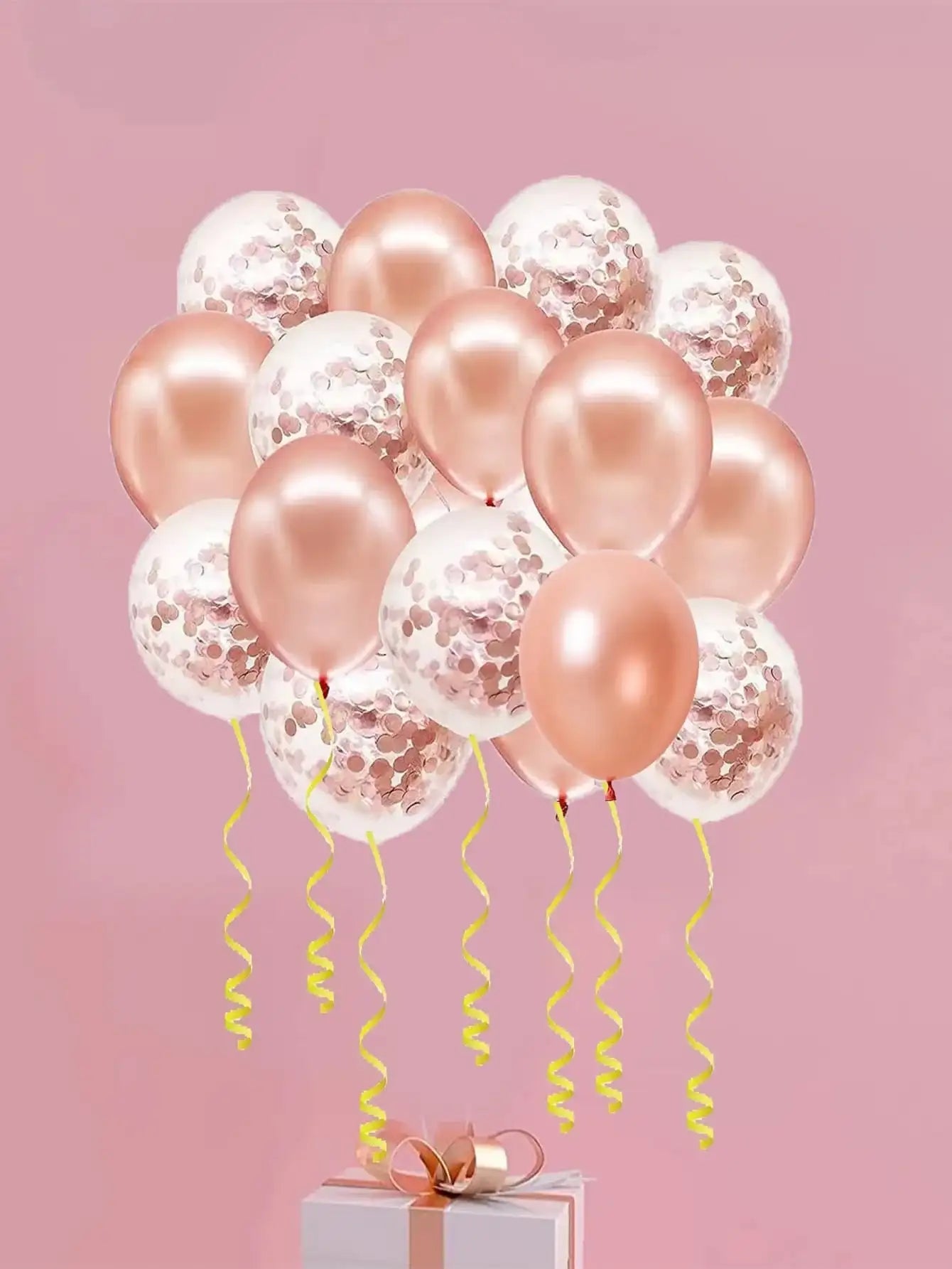 Un amas de ballons en latex rose gold et transparents remplis de confettis avec des rubans dorés, disposés au-dessus d'une boîte cadeau.